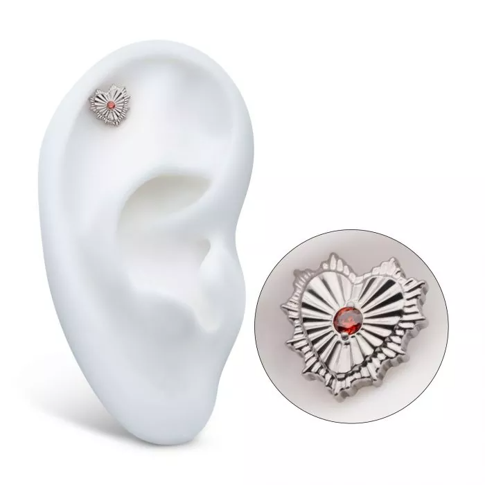 A silver heart-shaped stud earring with a red gemstone in the center, displayed in a model ear with a separate close-up shot.